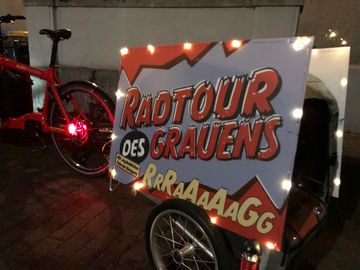ein Plakat auf einem Fahrrad-Anhänger. Auf dem Plakat steht: "Radtour des Grauens". Das Plakat ist ringsum von kleinen Lämpchen beleuchtetin Moers