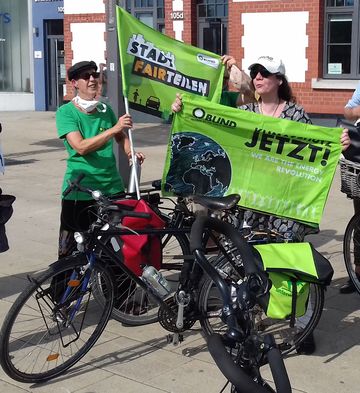 zwei Personen stehen hinter ihren Fahrrädern und hslten Fahnen in der Hand. Auf einer Fahne steht "Stadt Fairteilen", auf der anderen "Klimaschutz jetzt"