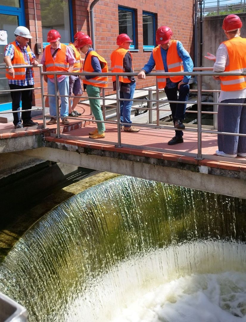 Mehrere Personen mit Schutzhelmen und Warnwesten stehen auf einem Steg über einem runden Becken in einer Kläranlage, in das von allen Seiten Wasser hineinläuft.